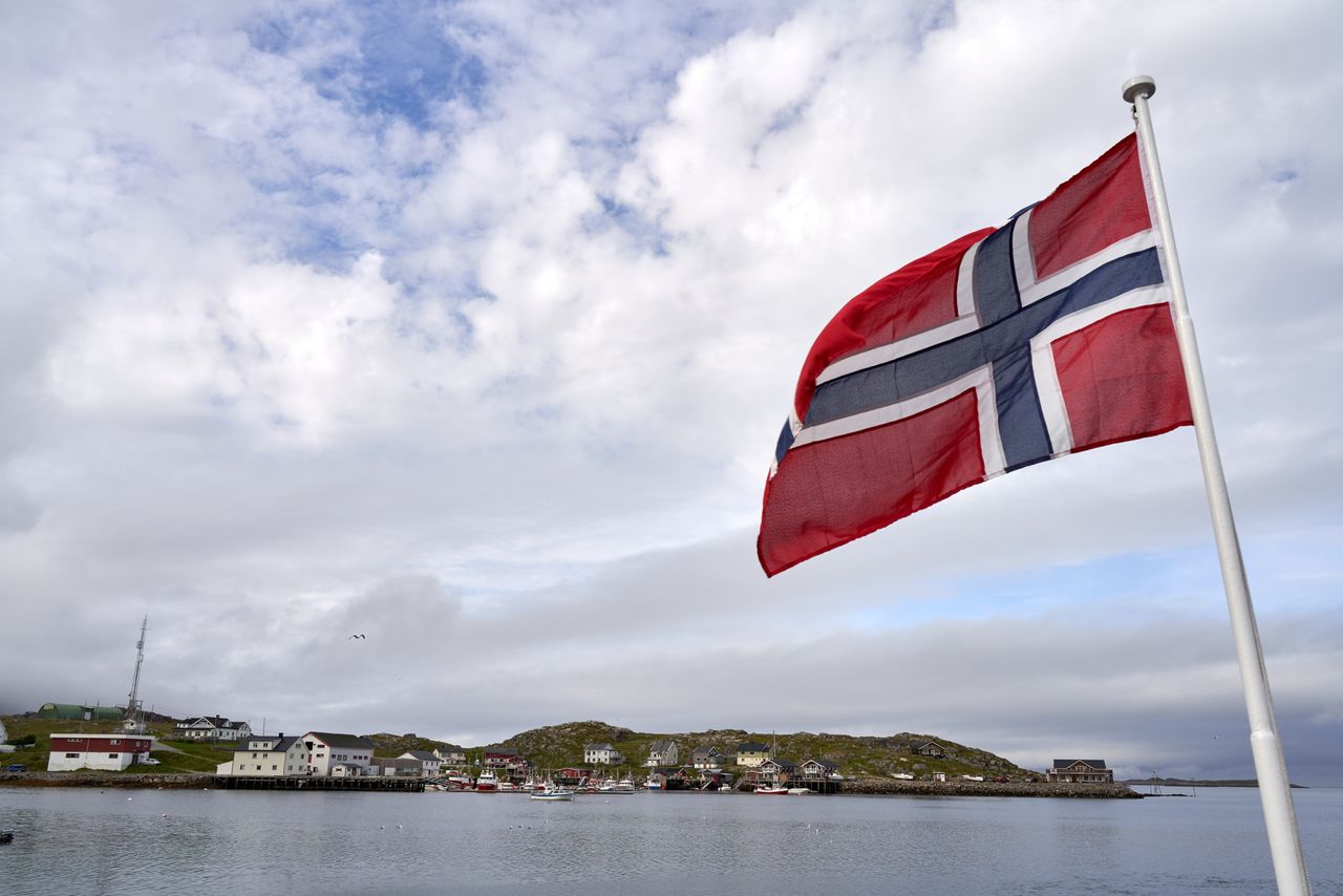 Norwegian flag waving by a serene coastal village on a cloudy day with calm waters.